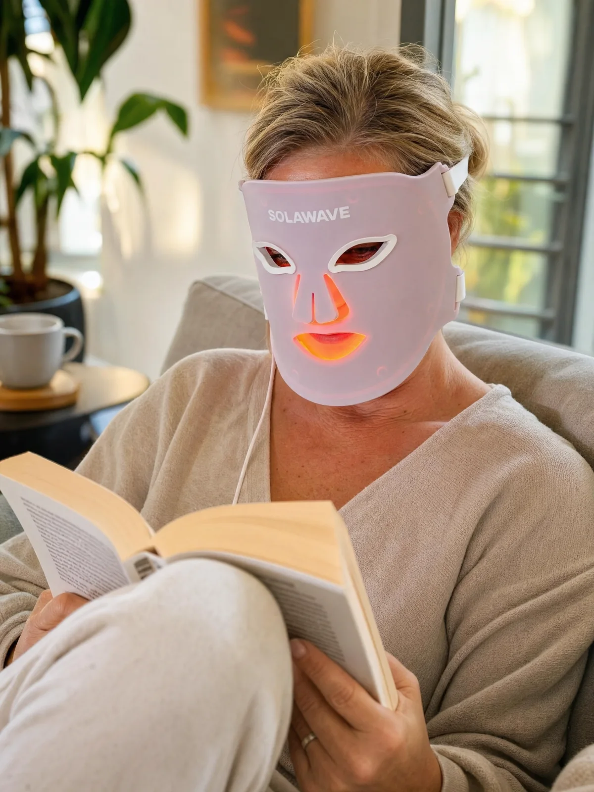 A woman in her fifties reading on a sofa while wearing the pink Solawave Wrinkle Retreat Pro LED mask; editorial illustration