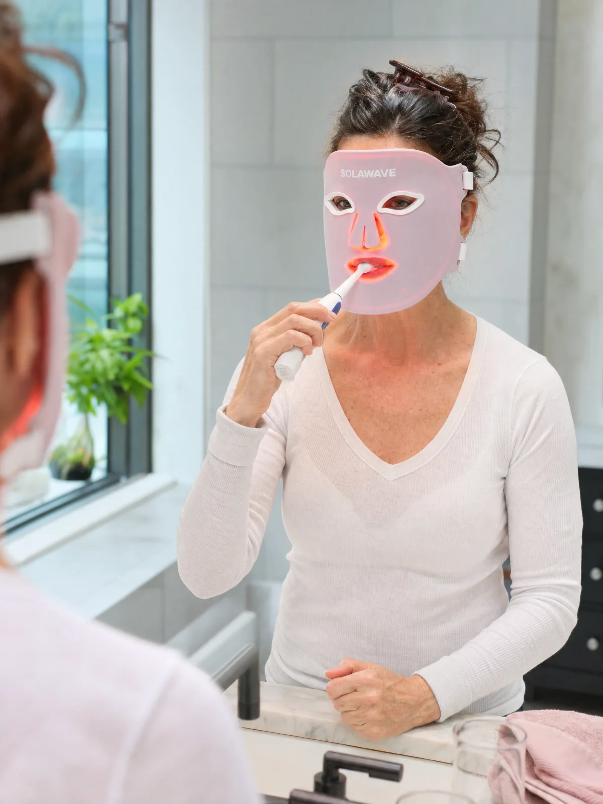 A woman brushing her teeth at a bathroom sink while wearing the Solawave LED mask; editorial illustration