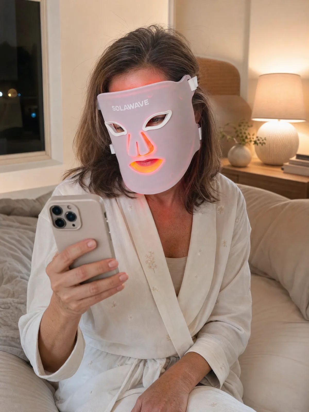 A woman sitting on her bed at night checking her phone while wearing the Solawave LED mask; editorial illustration