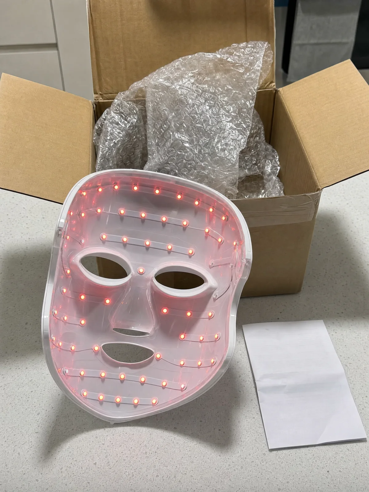 A generic rigid plastic LED mask with sparse bulbs lying in an opened cardboard shipping box; editorial illustration