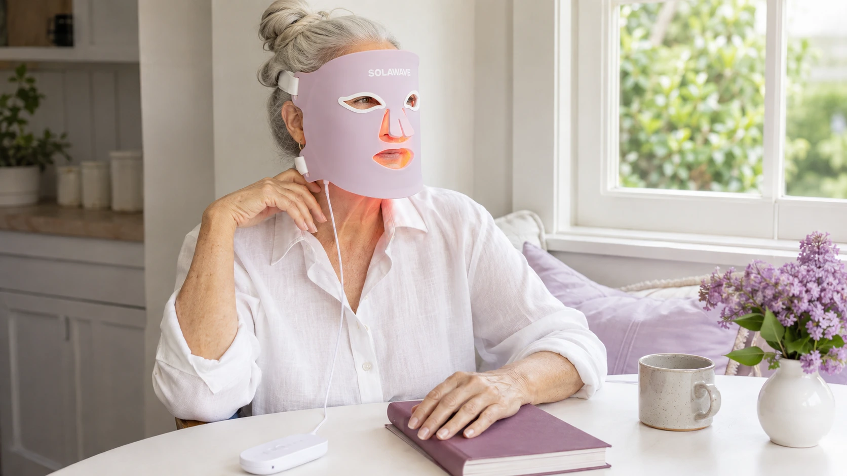Editorial illustration of a woman in her sixties wearing a Solawave mask at home with the controller beside a book